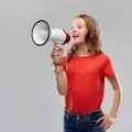 communication, feminism and people concept - smiling teenage girl in red t-shirt speaking to megaphone over grey background